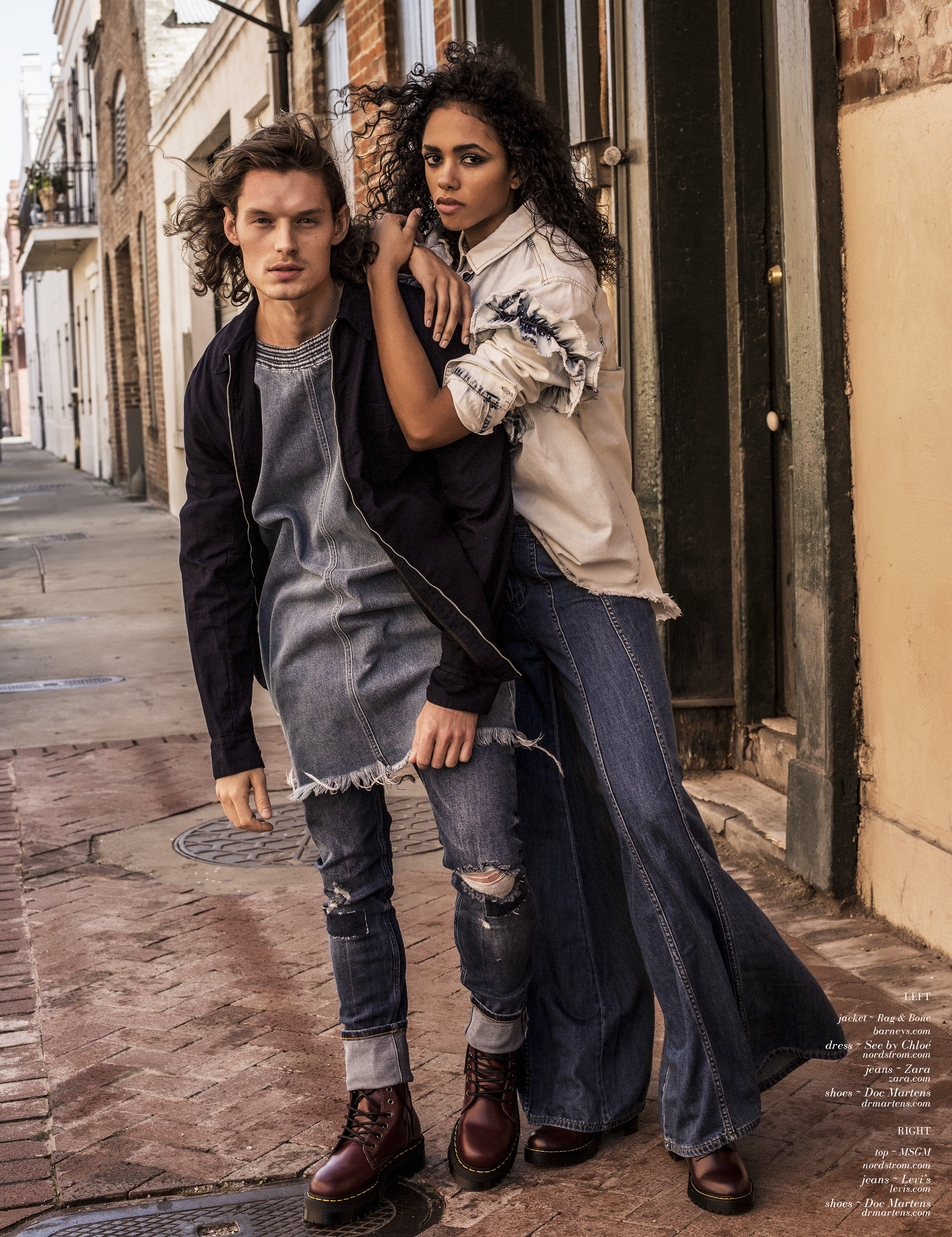 Two fashion models in denim and casual wear pose outdoors on a brick-paved street with buildings in the background, captured in medium shot.