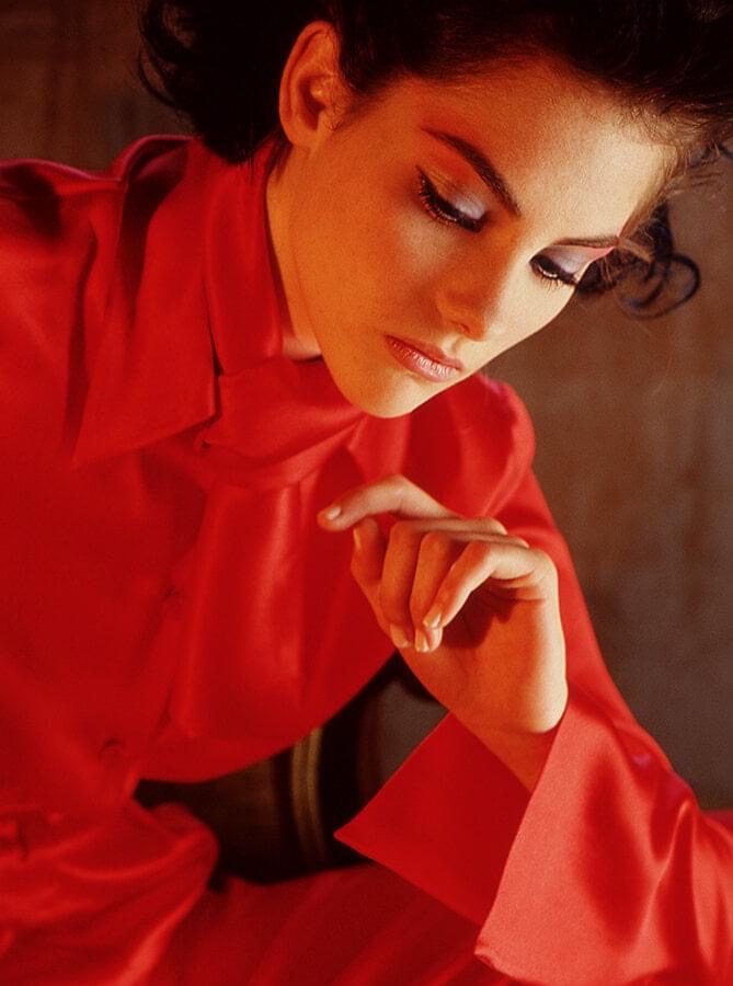 Portrait of an adult woman in a red silk blouse, posed with hand near her face. Soft, warm lighting with a dark background.
