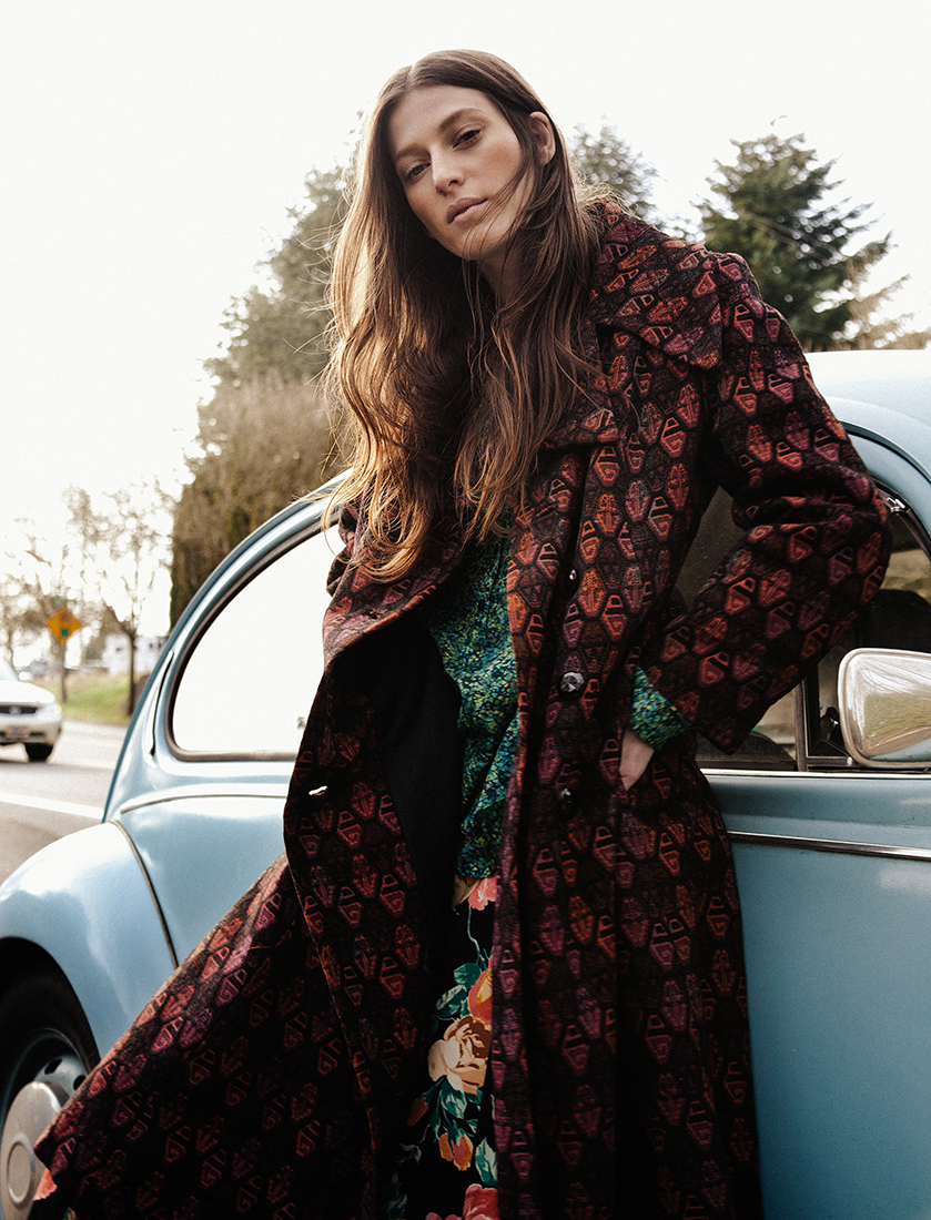 A woman in a patterned coat stands next to a vintage blue Volkswagen Beetle with trees and another car in the background on a sunny day.