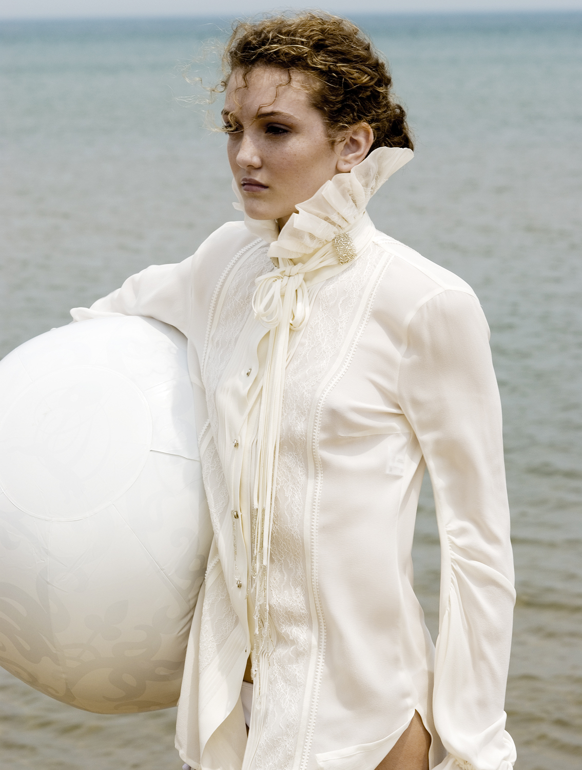 Young woman in a white, ruffled shirt holds a large white ball near the ocean, evoking a sense of elegance and serenity in an outdoor setting.