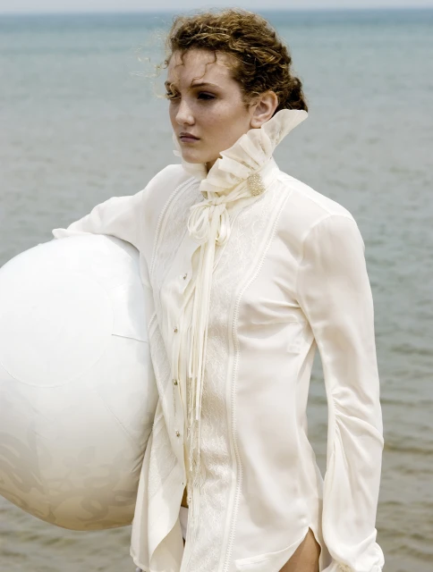 Young woman in a white, ruffled shirt holds a large white ball near the ocean, evoking a sense of elegance and serenity in an outdoor setting.