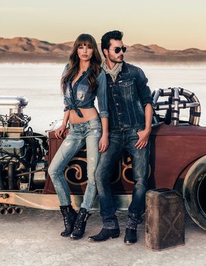 Fashion models in denim clothing pose next to a vintage hot rod against a desert landscape at sunset. Denim outfits and goggles.