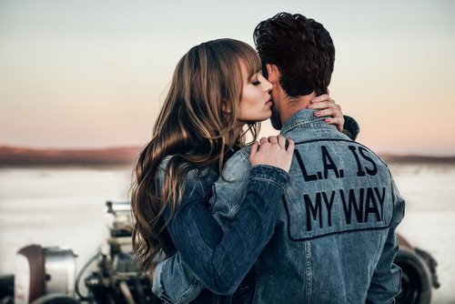 Romantic couple embracing outdoors at sunset. Woman kissing man wearing denim jacket with 'L.A. IS MY WAY' text. Serene landscape backdrop.