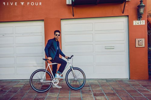 A man in a blue jacket and jeans rides a white bicycle in front of a white garage door with an orange wall in the background.