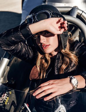 Adult female model in black bomber jacket and aviator helmet posing in vintage racecar with silver watch, eye-level angle, outdoor setting.
