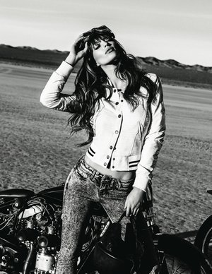 Monochrome portrait of a young adult woman with long hair wearing a jacket and jeans, posing next to a motorcycle in the desert.