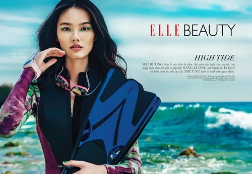 Asian woman in wetsuit poses on beach with blue water fins near Elle Beauty ad, wearing makeup and a pink smartwatch outdoors.
