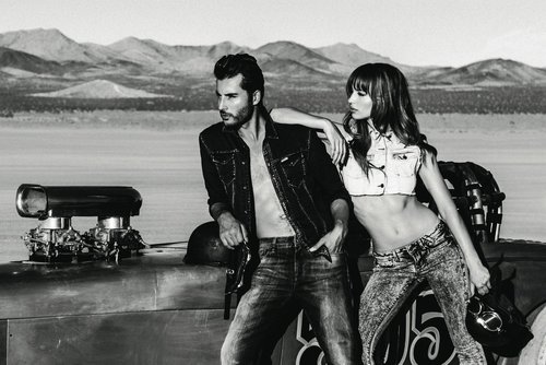 Black and white image of a woman and man posing next to a vehicle in a desert landscape with mountains in the background.
