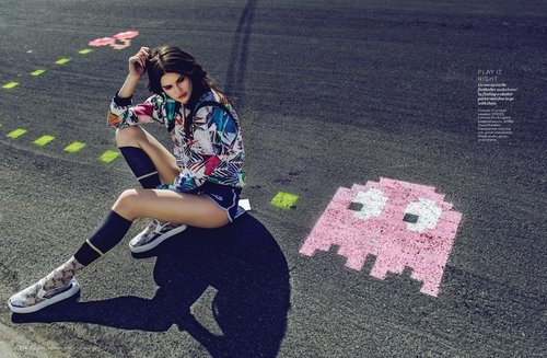 A woman in a colorful jacket sits on asphalt with pink game character graphics, wearing knee-high socks and athletic shoes, outdoors.