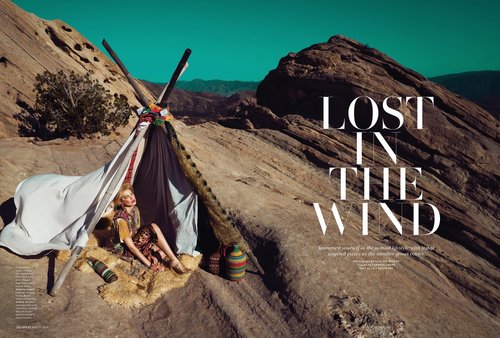 A woman sits inside a teepee made of sticks and cloth, located in a desert setting with large rocks. Fashion editorial photography.