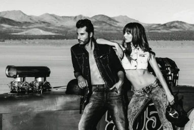 Man and woman posing outdoors next to a vintage hot rod vehicle in a desert landscape shot in black and white.