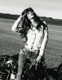 Monochrome photograph of a woman leaning against a motorcycle in a desert landscape, wearing a buttoned jacket and patterned jeans.