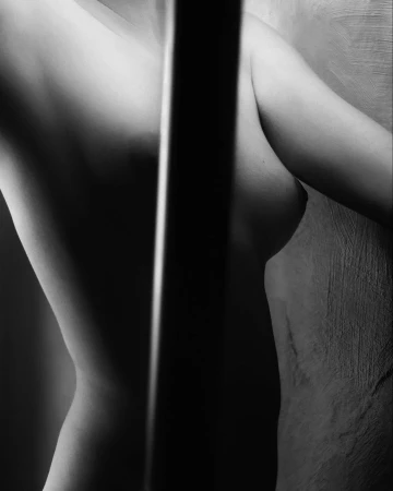 Monochrome torso study of a woman partially blocked by a vertical shadow bar against a textured wall by Omar Cruz.