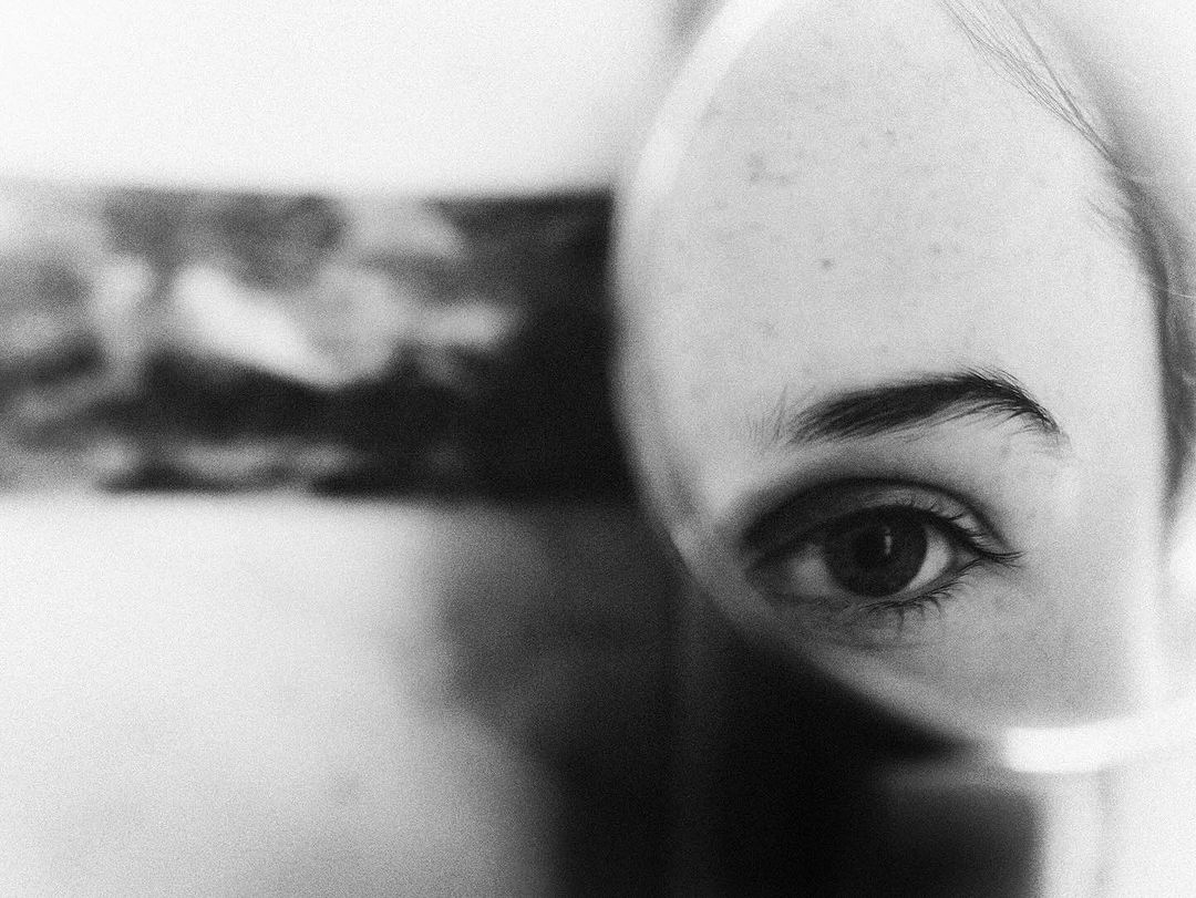 Monochrome close-up of an adult female's face, featuring a prominent eye, eyebrow, and blurred background, evoking an intimate and personal perspective.