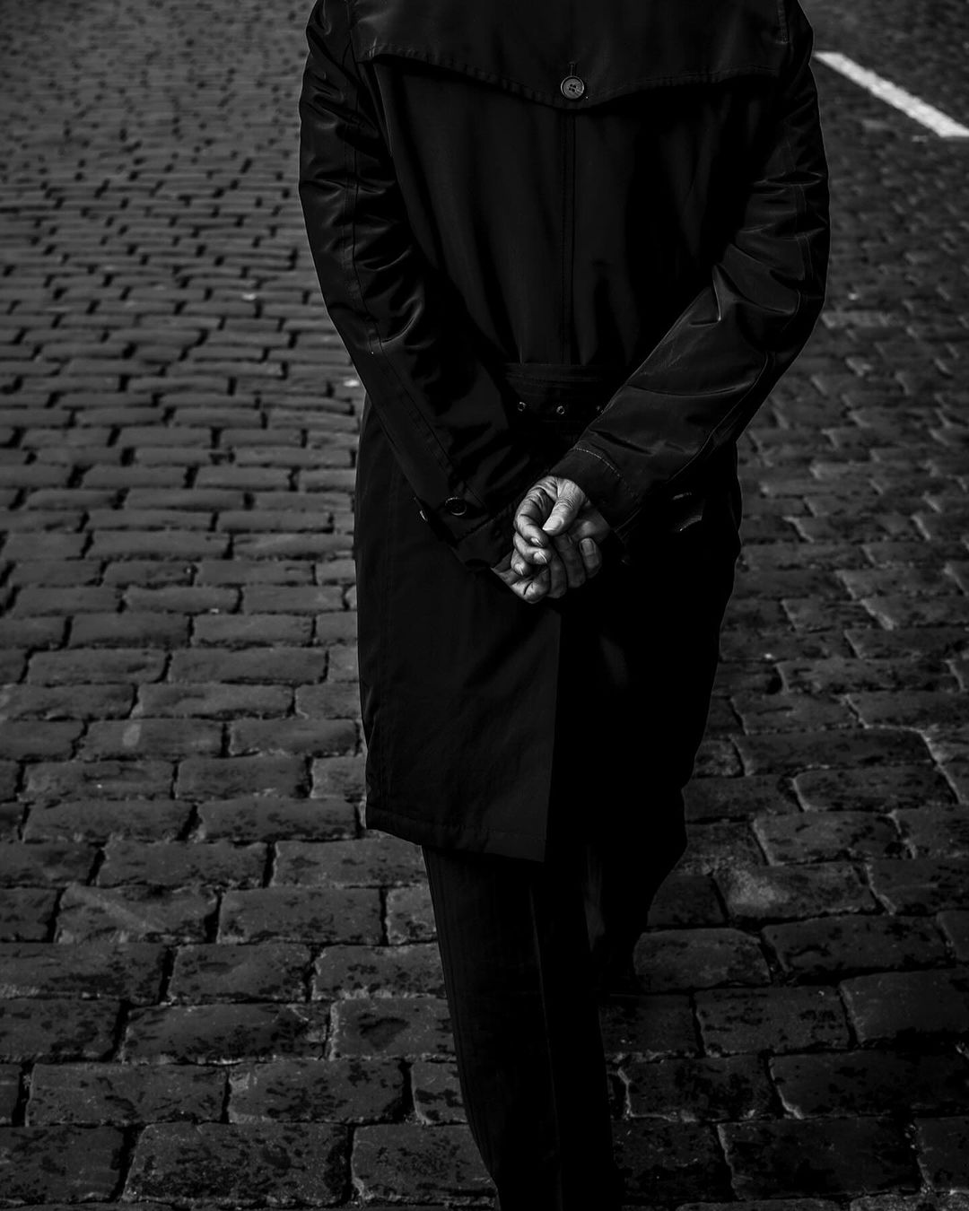 Black and white photo of a person in a dark coat with their hands clasped behind them, standing on a cobblestone street.