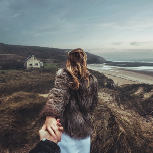 Woman in fur coat leads a person by the hand on a grassy dune overlooking a beach with a house in the distance under a cloudy sky.