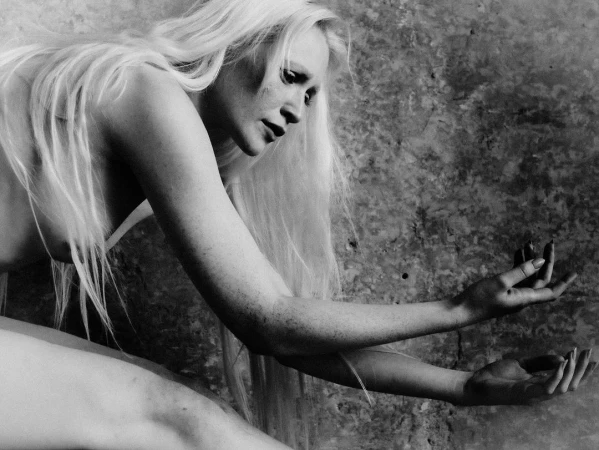 Black and white portrait of a blonde woman with long hair holding out her hands against a textured concrete wall.