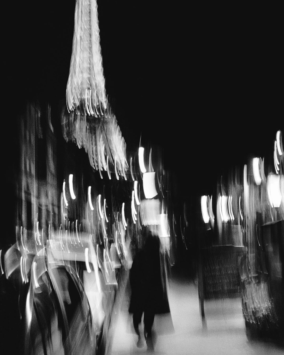 Abstract black and white view of the Eiffel Tower at night, with the blurred figure of a person walking along the street.