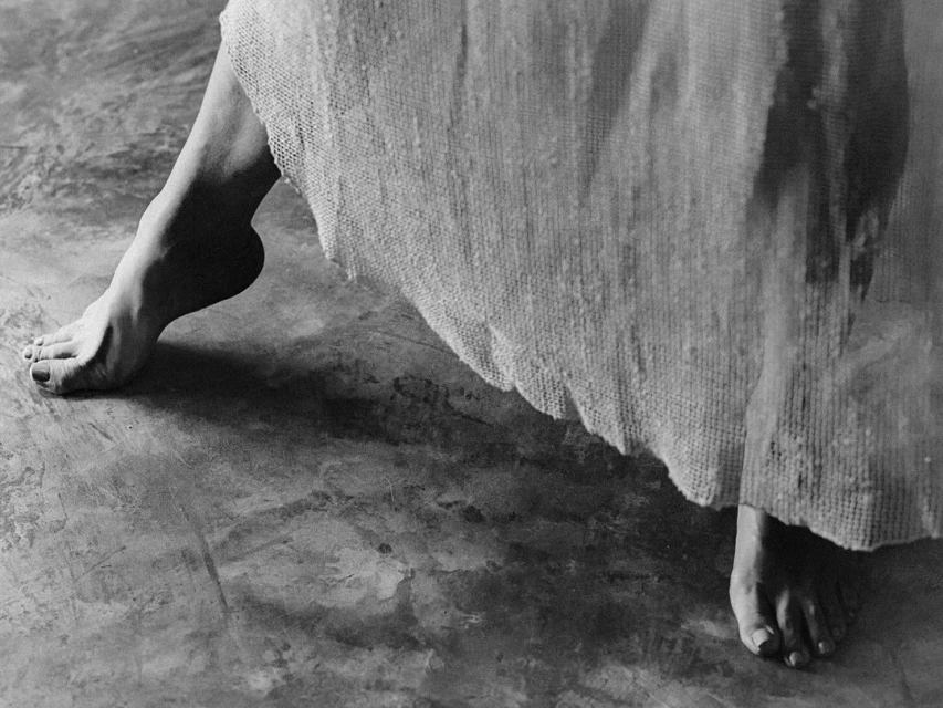 Close-up, black and white image of a person's bare feet standing on a textured surface, with a sheer white fabric draped nearby.