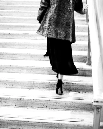 Monochrome rear view of a person walking up marble stairs wearing a textured coat and heels.