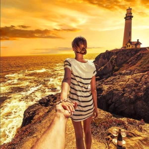 A woman in a striped shirt reaches for a man's hand on a rocky cliff overlooking the ocean with a lighthouse in the background at sunset.