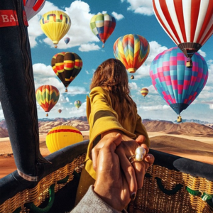 Woman in yellow coat holds hand of another person as they view hot air balloons in a desert landscape with a bright blue sky.