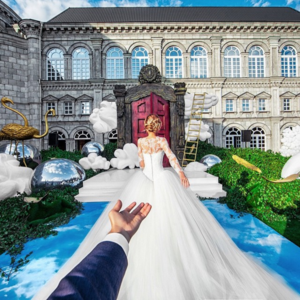 A bride in a white dress is reaching towards a hand in front of a building with a stage installation, featuring clouds and water.