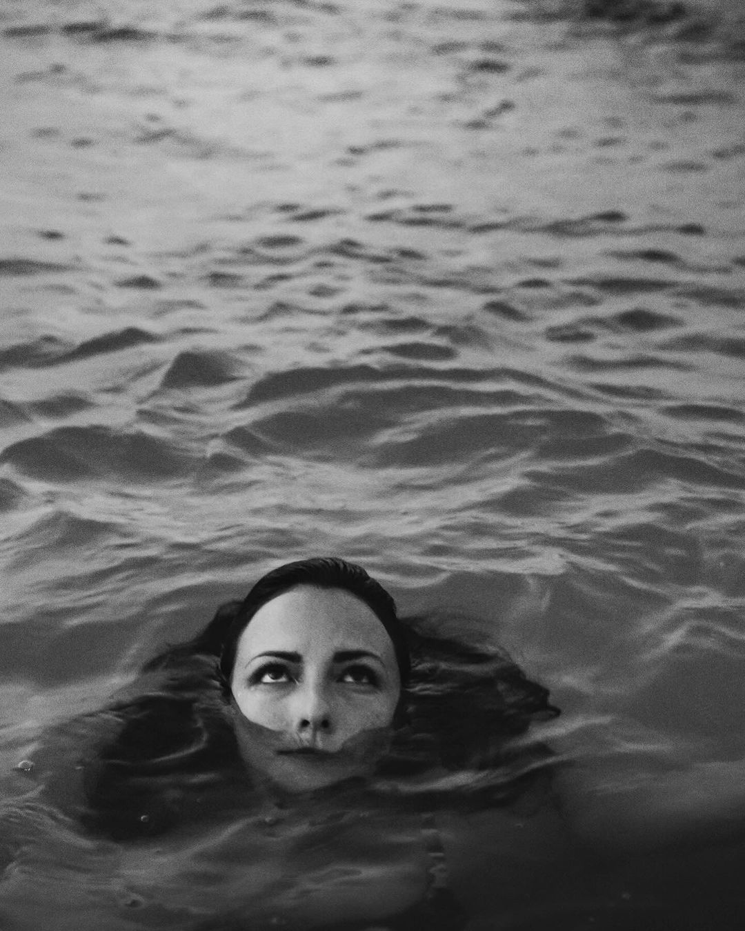 Monochrome portrait of a woman submerged in water, looking upward with ripples surrounding her head.