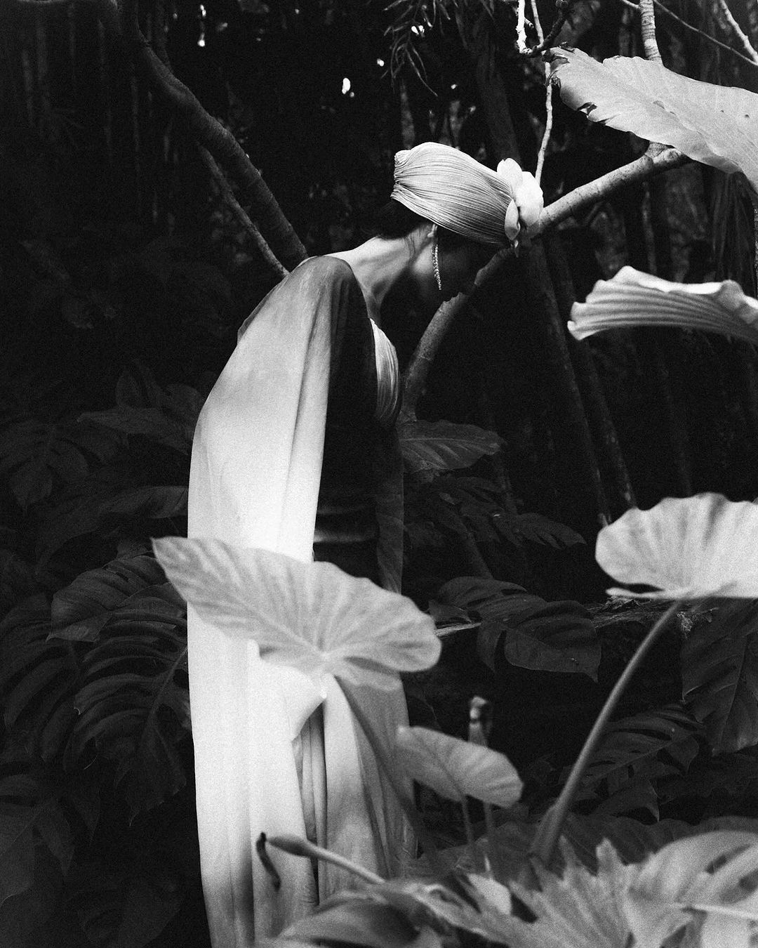 Black and white photograph of a woman with a head covering standing in a tropical garden surrounded by large leaf plants and trees.