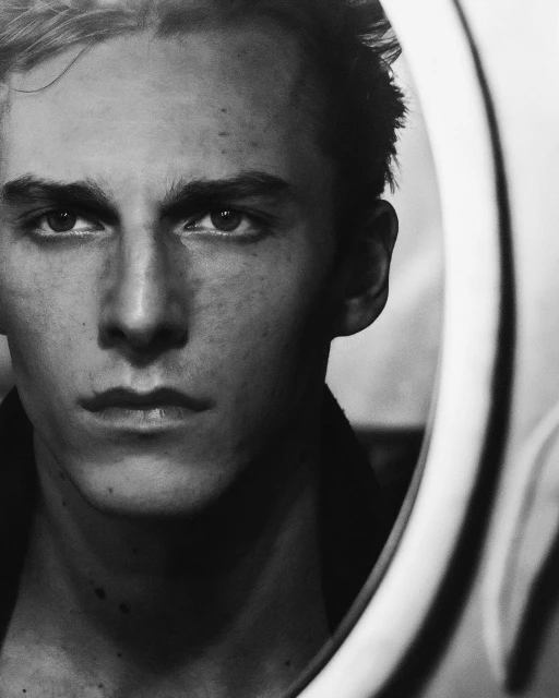 Monochrome portrait of an adult male with dark hair and light skin, reflected in a mirror with a neutral expression. 