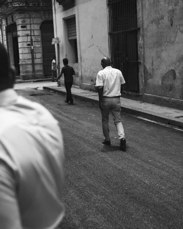 Black and white street photograph of a bald man walking away down a narrow urban sidewalk and asphalt road by Omar Cruz