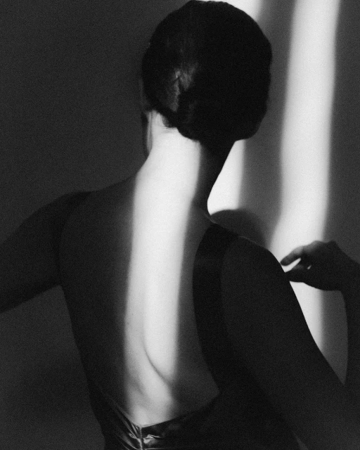 Monochrome image of a woman's bare back highlighted by a vertical beam of light, set against a shadowed wall in a minimalist style.