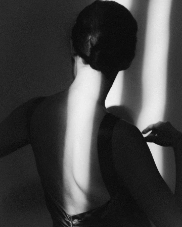 Monochrome image of a woman's bare back highlighted by a vertical beam of light, set against a shadowed wall in a minimalist style.