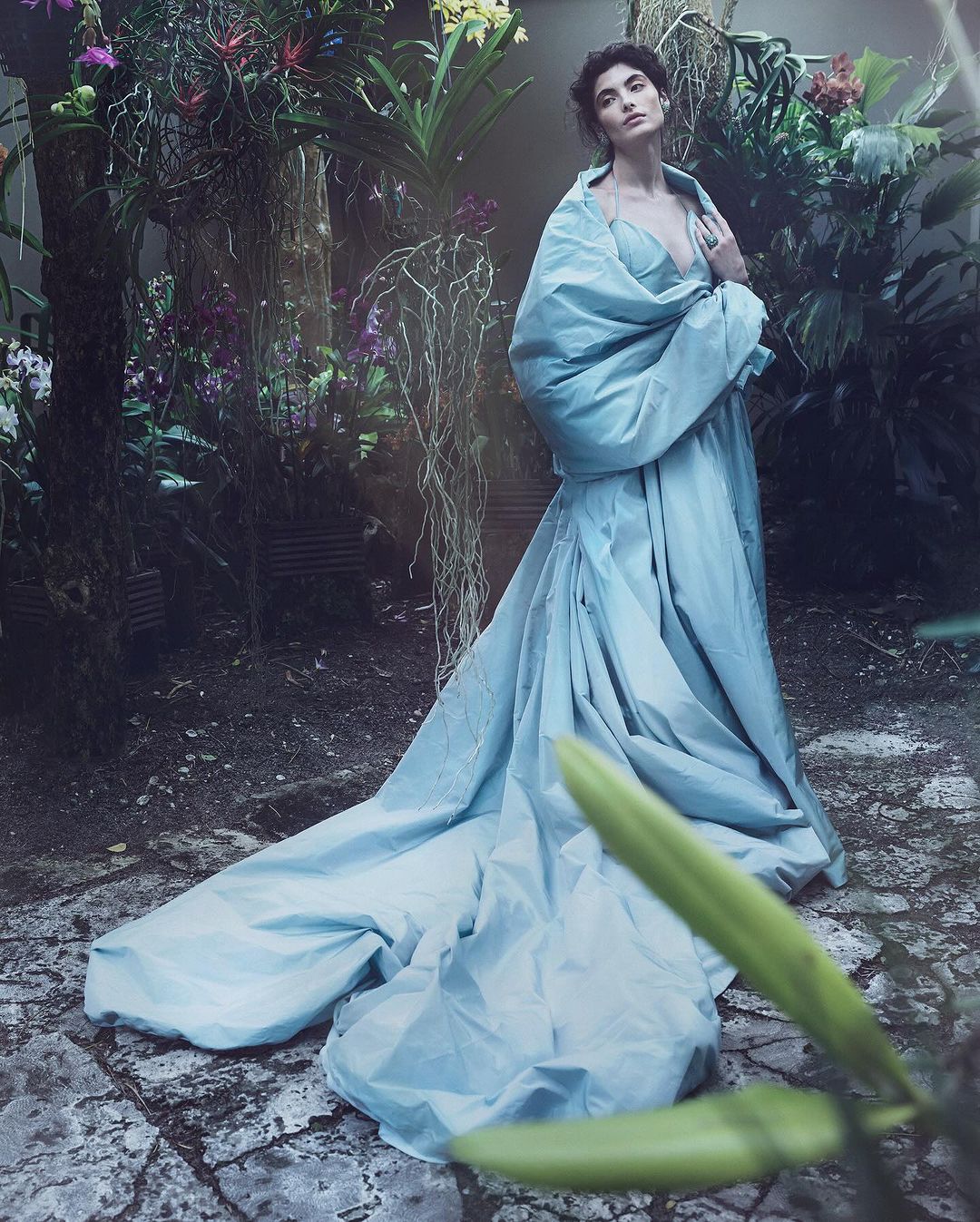 Woman in flowing blue gown stands amid lush botanical garden with orchids and ferns, fashion editorial photography.