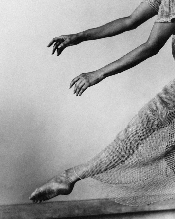 Monochrome art photo of dancer's leg and foot extended, hands reaching up, wearing textured mesh dress, against pale background.