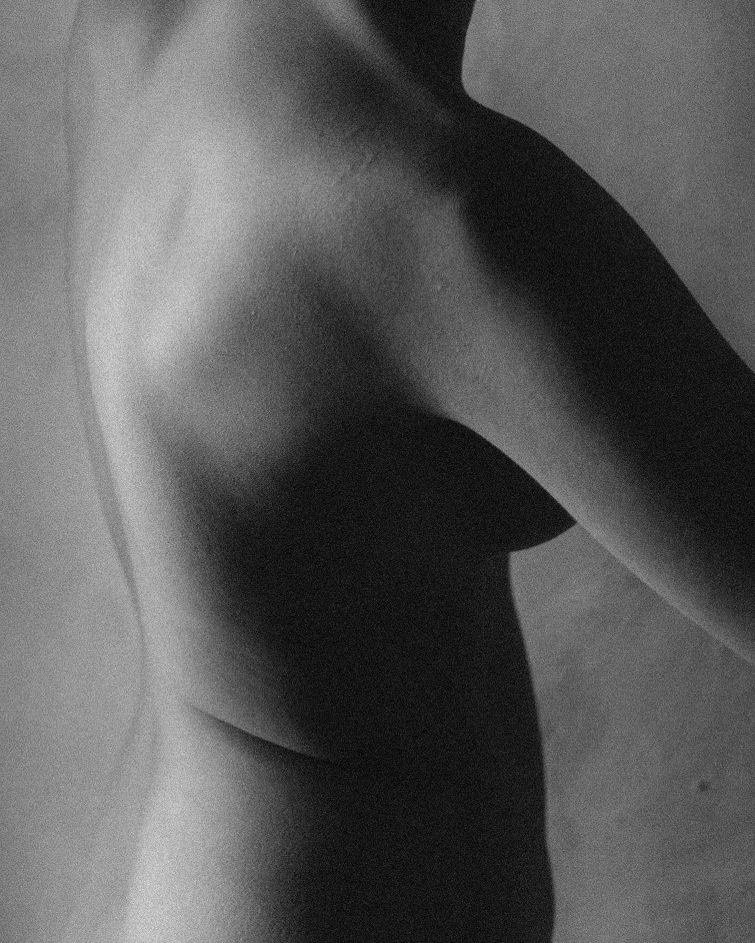 Monochrome nude upper body image of an adult female with visible arm, shoulder, and chest against a neutral background with soft, natural lighting.
