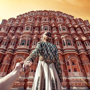 Woman in traditional Indian dress is holding a hand out toward Hawa Mahal palace in Jaipur, India, under a bright orange sky.