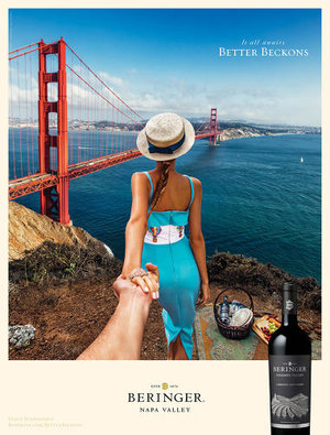 Woman in hat, light blue dress leads hand to Golden Gate Bridge view from grassy cliff with picnic basket, bottle of Beringer wine.