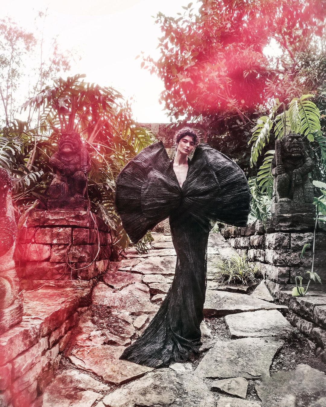 Fashion model wearing a sculptural black gown in a lush jungle setting with ancient stone architecture and diffused lighting.
