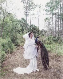 Model in flowing white gown with hooded cape holding a peacock outdoors on a forest path by Omar Cruz