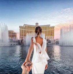 Woman in a white dress leading someone by the hand in front of water fountains and a large grand hotel building at sunset.