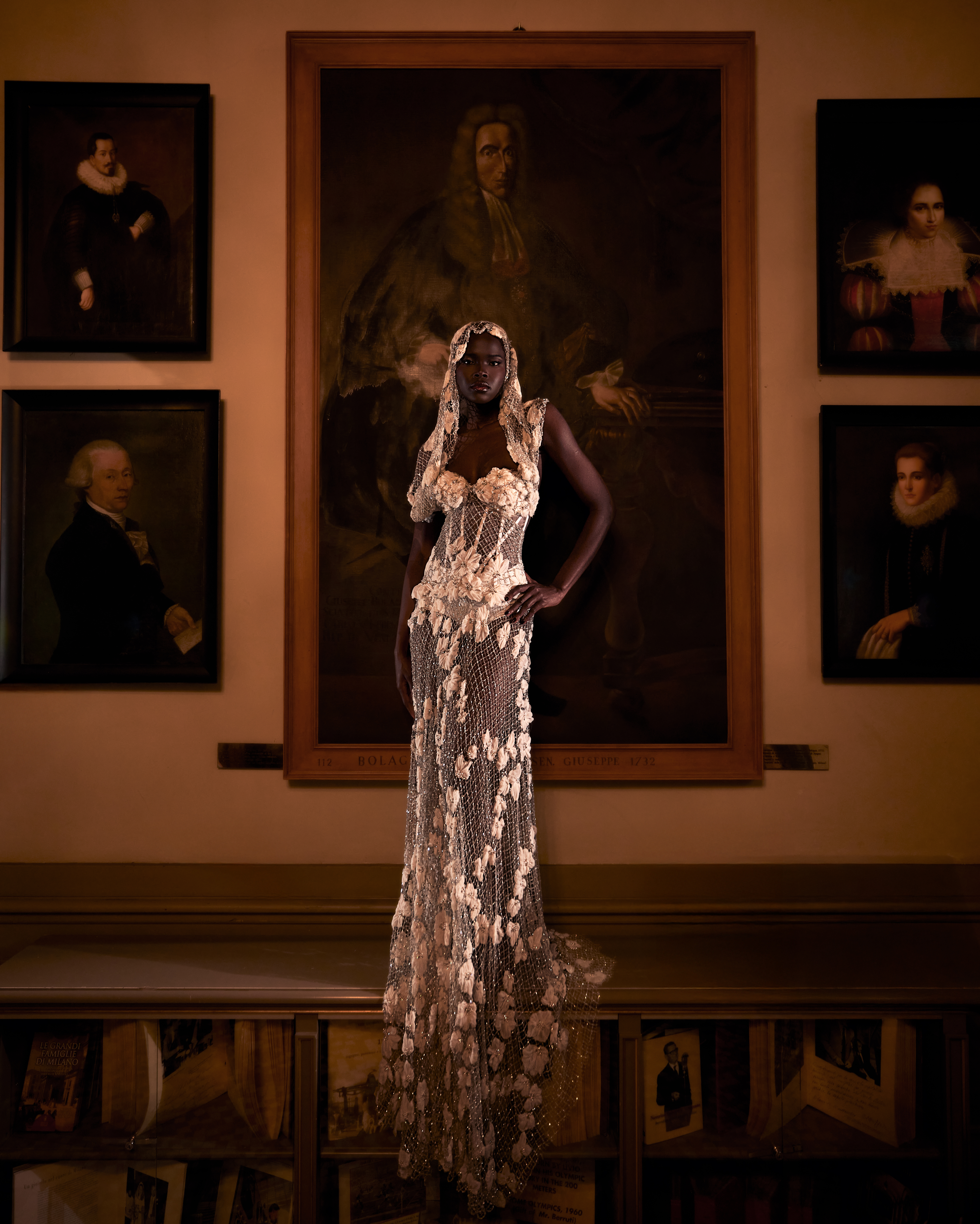Fashion shot of an African woman wearing a white lace gown posing indoors in front of antique portraits hanging on a wall.