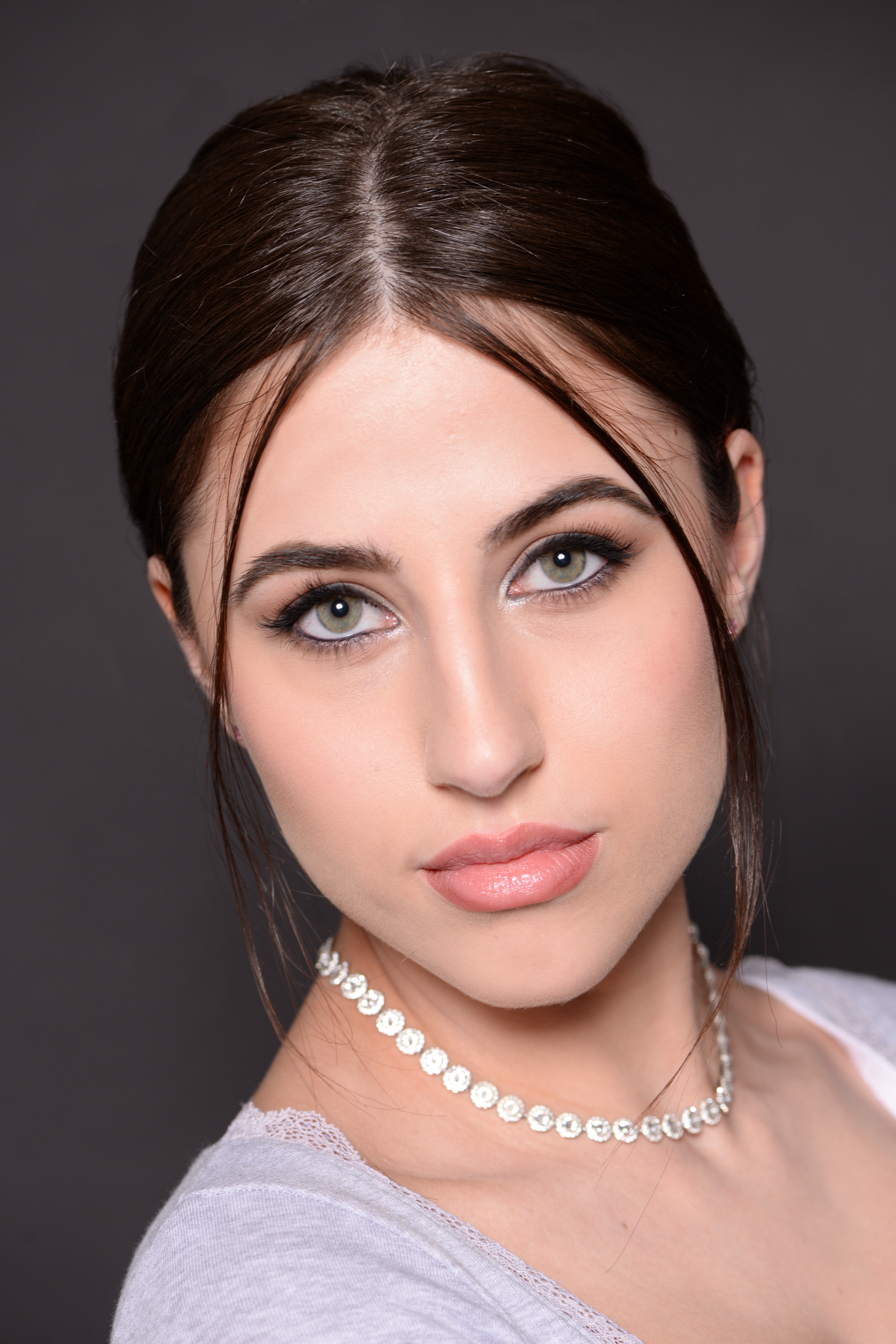 Close-up portrait of a woman with dark hair and grey eyes wearing a pearl necklace against a dark grey studio background.