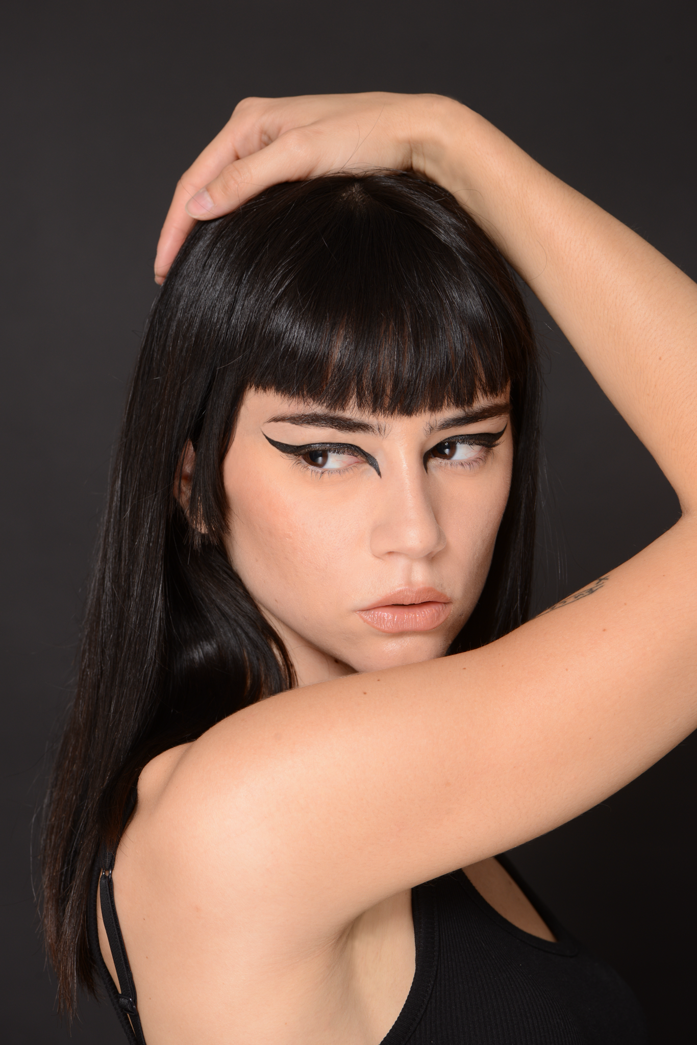 Close-up portrait of a brunette woman in a black top with dramatic eyeliner, holding her hand on her head against a dark gray background.