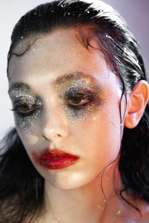 Close-up portrait of a young woman with wet black hair and dramatic glitter makeup on her eyes and red lips, against a soft gradient backdrop.