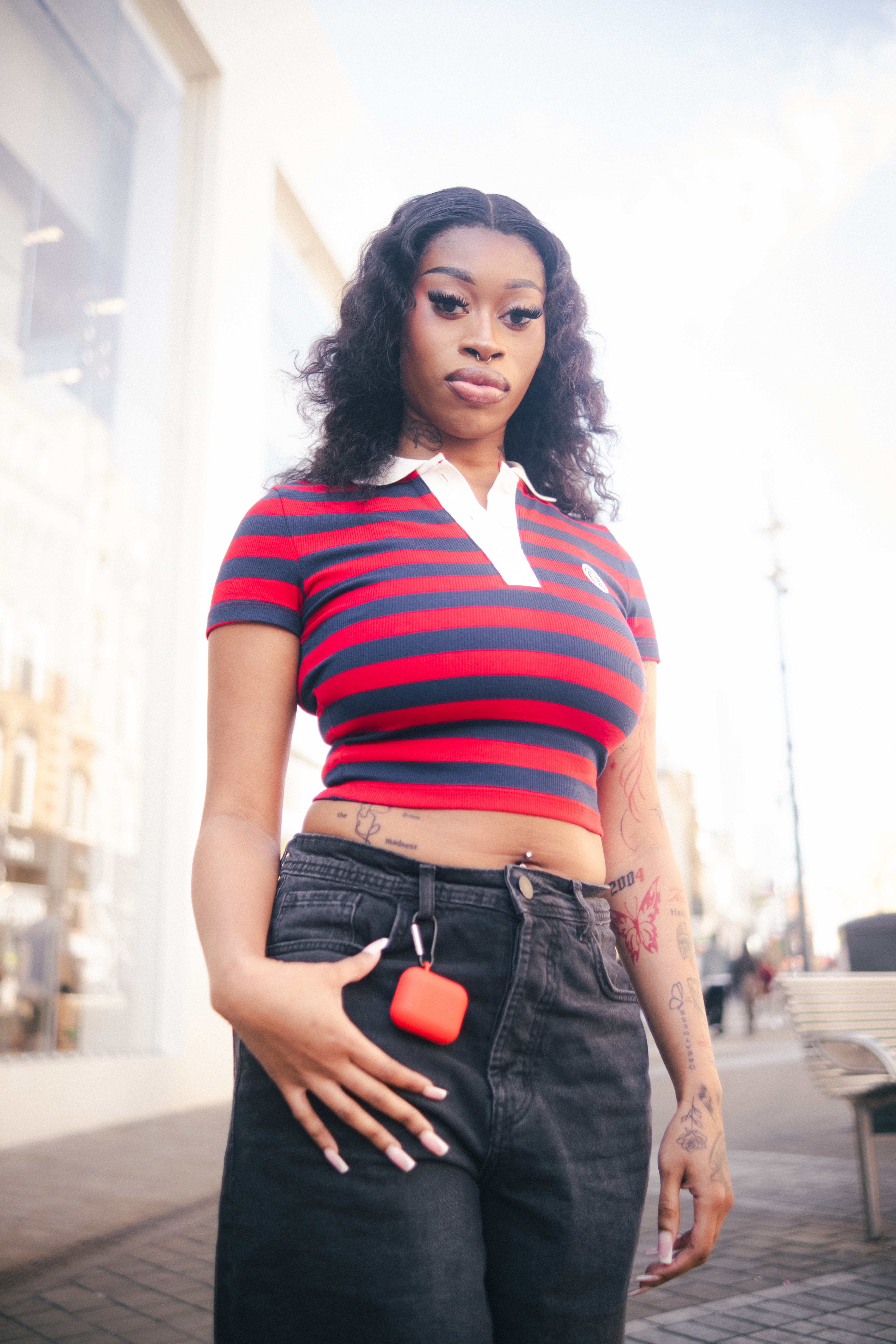 A young Black woman stands outside wearing a red and blue striped shirt and black jeans with an orange case attached to her jeans.