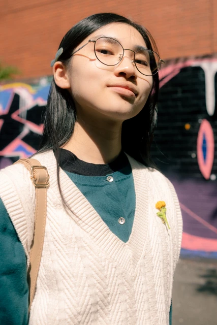 Eye-level shot of an Asian young adult woman wearing glasses and a sweater vest against a brick wall with colorful graffiti.