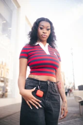 Young Black woman wearing a red and navy striped cropped polo shirt and black jeans outdoors on a city sidewalk.
