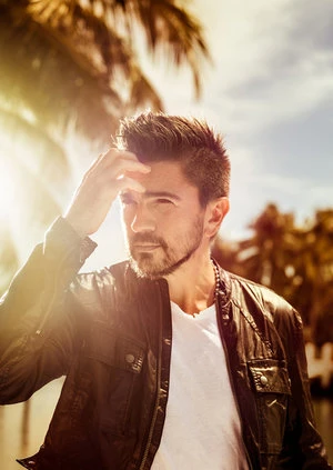 Handsome man in black leather jacket poses outdoors with palm trees and a sunny sky in background, wearing a white t-shirt.