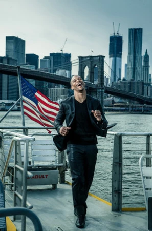 Musician Maury Hechavarria laughing on a boat deck with an American flag and city skyline in the background.